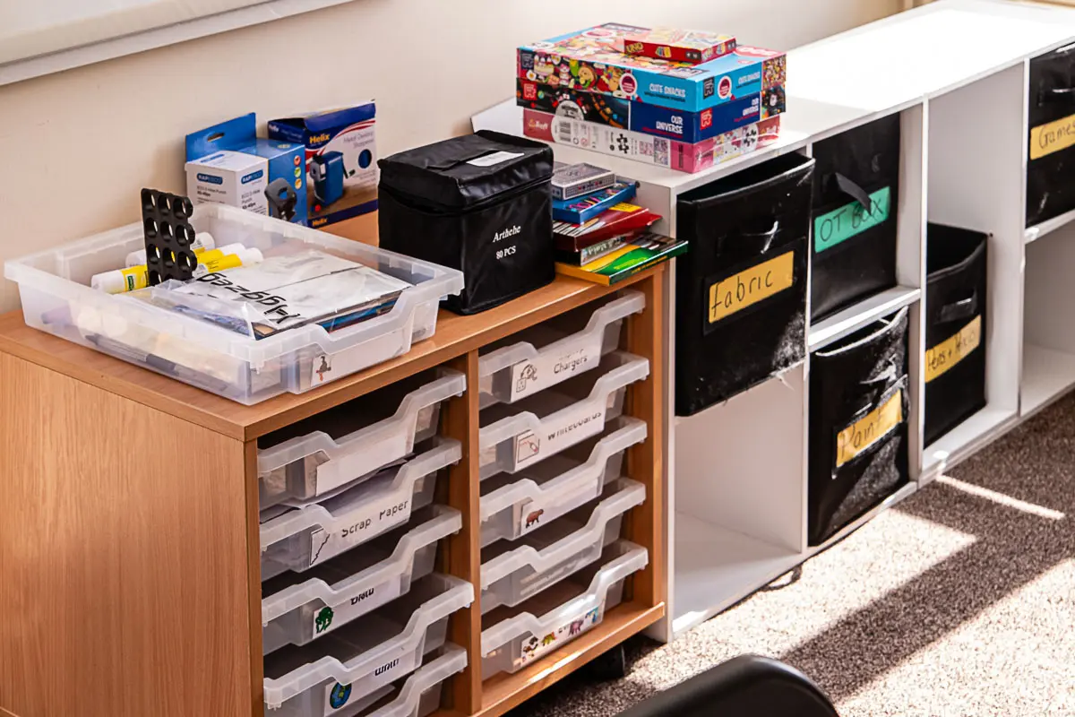 Organised shelves with plastic drawers and labelled black boxes holding school supplies and games.