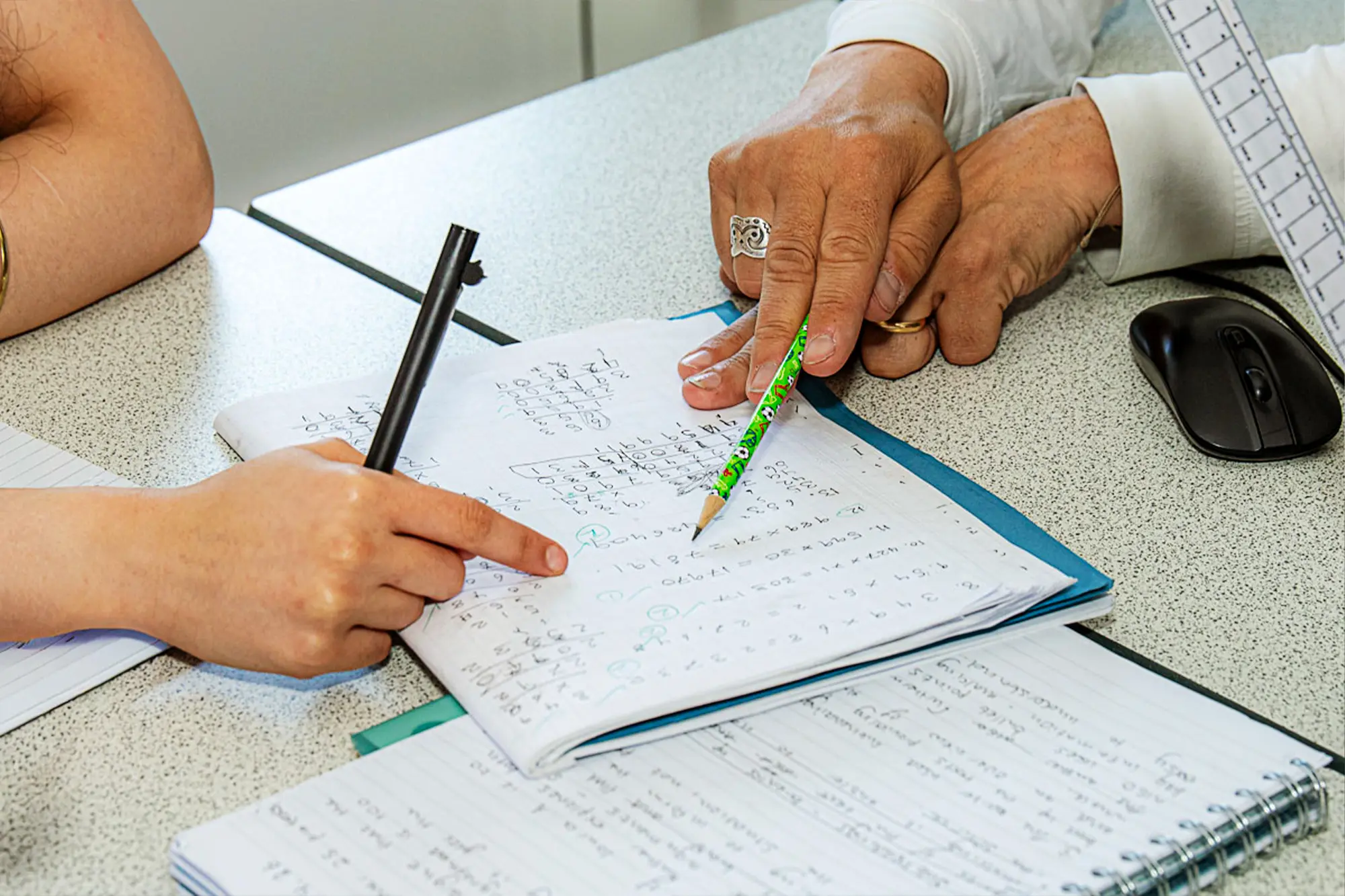 Hands of an Egham Park School teacher and student pointing at math problems in a notebook on a table during a study session.